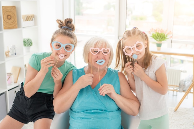 Smiling girls and senior woman with paper glasses and lips in light room