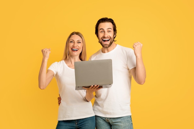 Smiling glad millennial caucasian guy and lady in white tshirts hold laptop making victory sign with hands