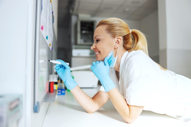 Assistente di laboratorio bionda femminile splendida sorridente che si appoggia sulla scrivania e che scrive sulla ricerca della lavagna bianca per il vaccino