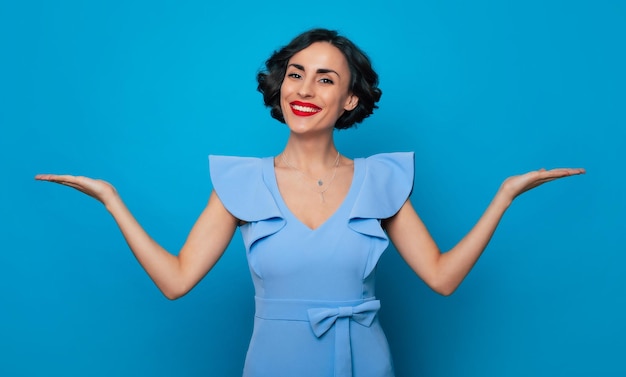 Smiling gorgeous young brunette woman with wavy hairstyle and bright toothy smile while she shows her empty palms in dress on blue background