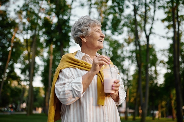 Smiling granny holds milk coctail in summer amusement park, back to childhood. Aged people lifestyle. Funny grandmother having fun outdoors, old female person outdoors