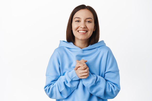 Sorridente donna grata si tiene per mano stretta, apprezza l'aiuto, grazie, sembra felice e grata, in piedi in felpa con cappuccio blu contro il muro bianco