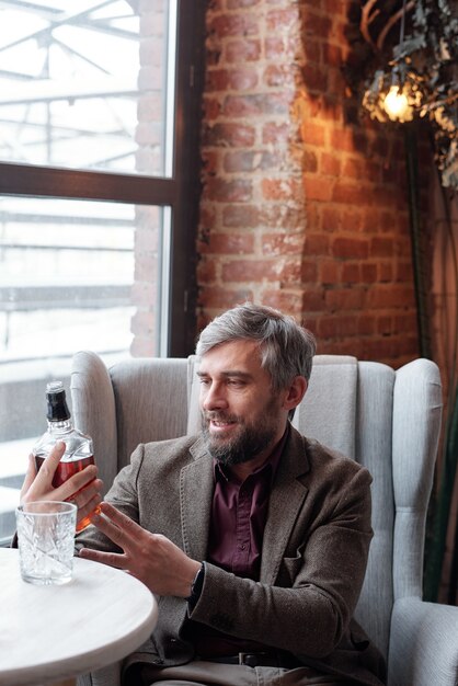 Smiling gray-haired businessman in brown jacket sitting at table and reading label of whisky bottle