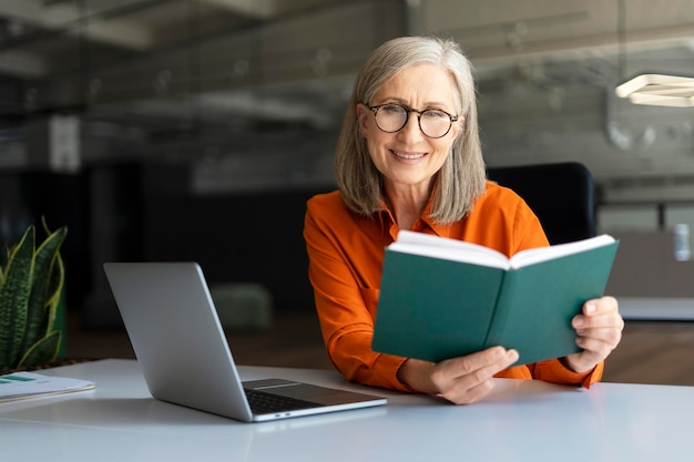 Sorridente direttore esecutivo della donna d'affari senior caucasica dai capelli grigi che indossa occhiali da vista controllando la sua agenda seduto alla scrivania con il computer portatile nell'interno di un ufficio moderno people business finance