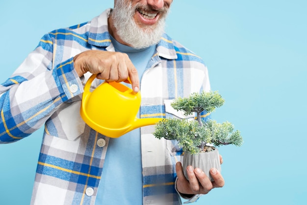 Smiling gray haired man wearing stylish shirt holding flower pot watering with yellow watering can