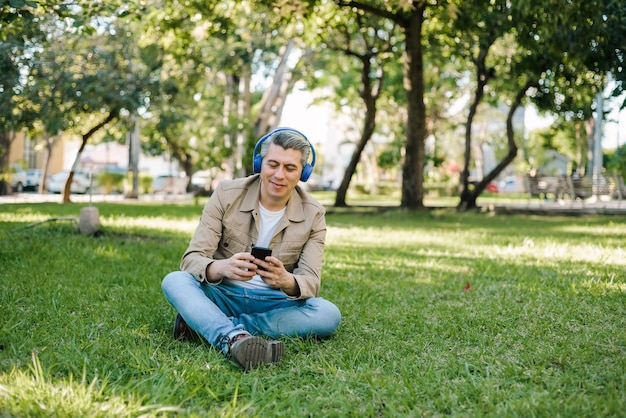 Smiling grayhaired man wearing headphones and typing on his cell phone sitting in the grass in a park