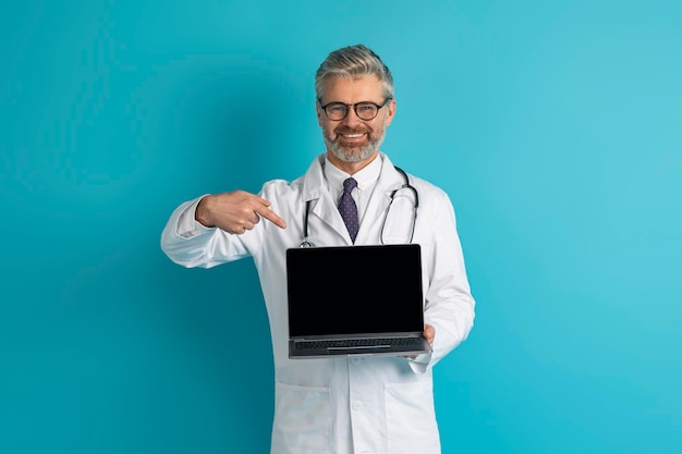 Улыбающийся седой кавказский врач показывает ноутбук с пустым экраном