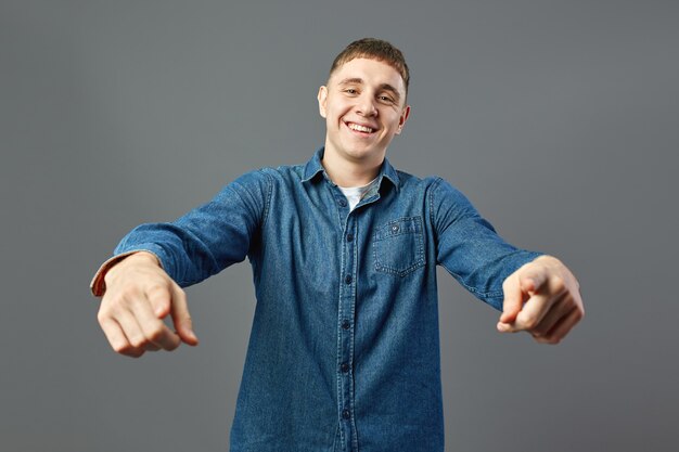 Smiling guy dressed in a jeans shirt points with his hands forward in the studio on the gray background .