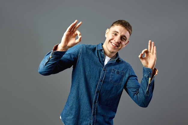 Smiling guy dressed in a jeans shirt shows with his fingers ok signs in the studio on the gray background .