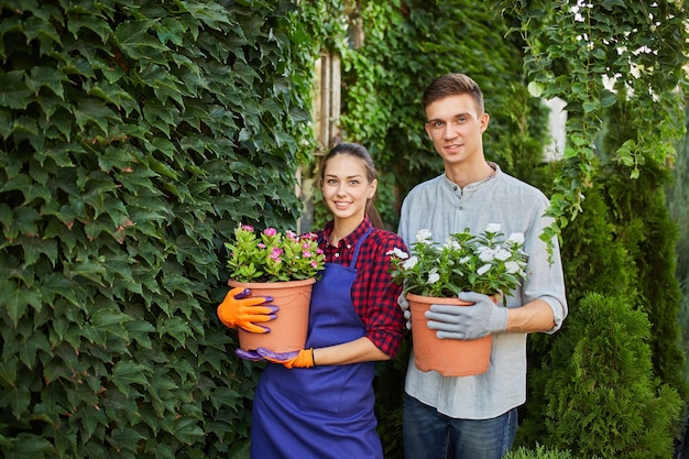 Smiling guy and girl gardeners are standing with pots with plants in their hands against a wall covered with green ivy in the beautiful garden on a sunny day .