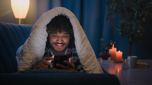 Smiling guy indian man arabian male at night evening home on couch under blanket cover with duvet