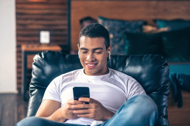 Smiling guy resting in an armchair, in headphones and with a smartphone, looking at the smartphone screen.