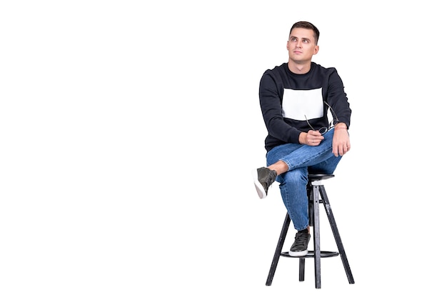 Smiling guy sits on high chair with leg on knee. White background and casual clothes. Copy space.