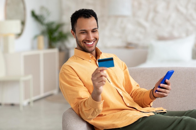 Smiling guy using phone and credit card at home