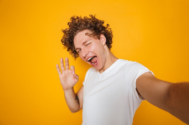 Smiling guy with brown hair showing ok sign on camera while photographing himself