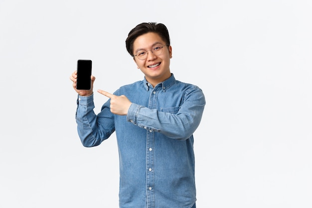 Smiling handsome asian guy with braces and glasses pointing finger at smartphone screen man showing ...