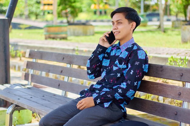 Smiling handsome Asian young man wearing batik sitting on park bench and talking on mobile phone People lifestyle concept