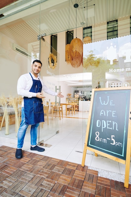 Smiling handsome bakery owner making welcoming gesture and inviting cusomers inside cafe