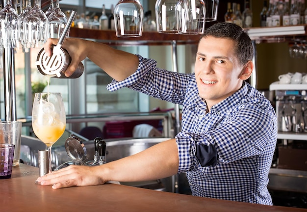Улыбающийся красивый бармен за барной стойкой наливает напиток в стакан из стального шейкера.