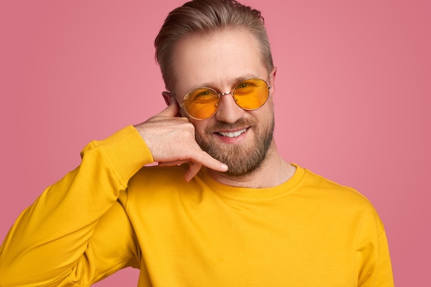 Smiling handsome bearded male in yellow sweatshirt and trendy sunglasses looking at camera and making call me gesture against pink background