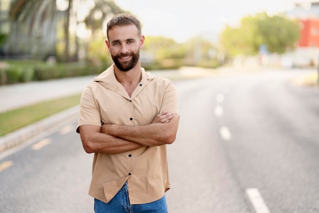 Smiling handsome bearded man with arms crossed looking at camera standing on the road, copy space