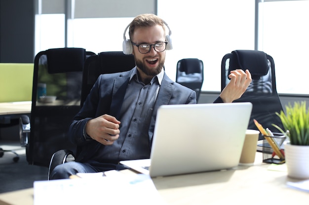 Smiling handsome businessman relaxing and listening music in earphones in a modern office.