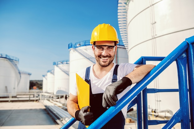 Smiling handsome caucasian worker in overall and with helmet on head leaning on the railing and holding folder with documents. oil production