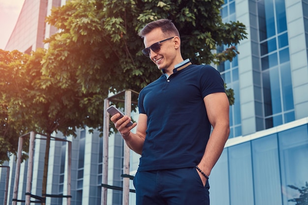 A smiling handsome fashionable man with a stylish haircut in sunglasses, dressed in a black t-shirt and pants, reads a message on the smartphone, stans in the modern city against a skyscraper.