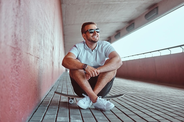 Smiling handsome fashionable skater guy in sunglasses dressed in a white shirt and shorts sitting on a skateboard under a bridge, looking away.