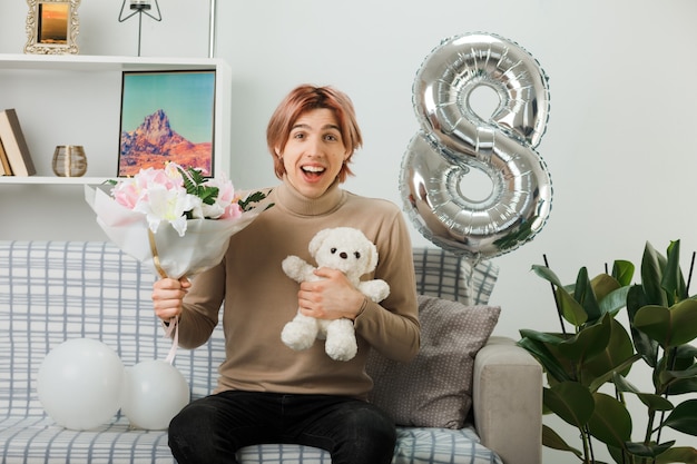 Smiling handsome guy on happy women day holding bouquet with teddy bear sitting on sofa in living room