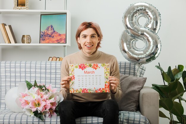 Smiling handsome guy on happy women day holding calendar sitting on sofa in living room