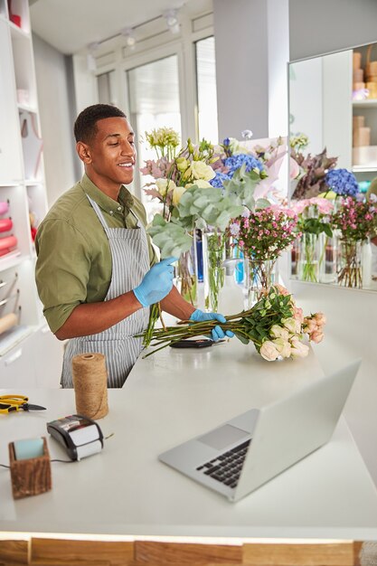 Smiling handsome male is making bouquet of roses while standing at counter with laptop and pos terminal