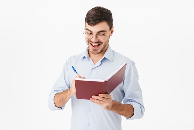 smiling handsome man in eyeglasses making notes in diary isolated over white wall