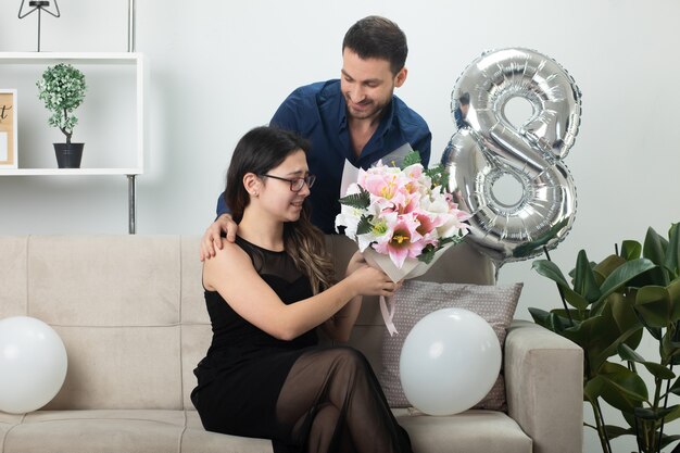 Smiling handsome man giving bouquet of flowers to excited pretty young woman in glasses sitting on couch in living room on march international women's day