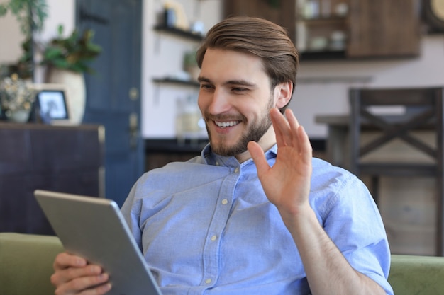 Smiling handsome man holding and using tablet with social networks at home, happy freelancer making online shopping, sitting on cozy sofa at living room