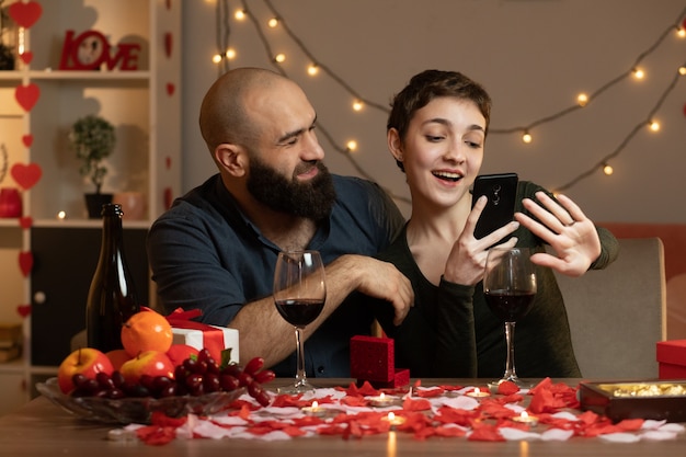 バレンタインデーにリビングルームのテーブルに座っている指で彼女の指輪の写真を撮って喜んできれいな女性を見て笑顔のハンサムな男
