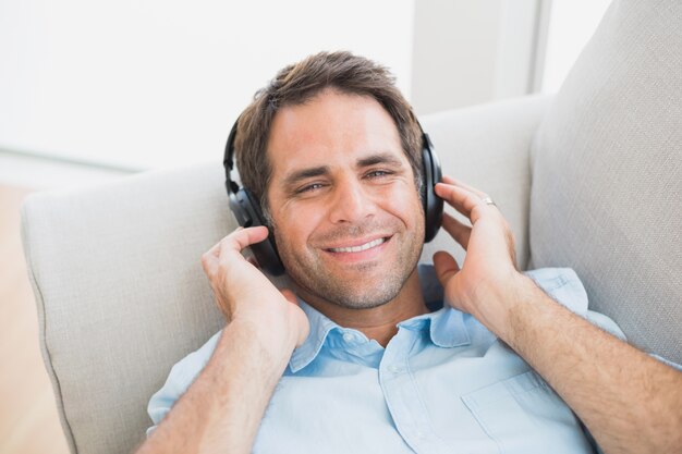 Smiling handsome man lying on sofa listening to music looking at camera