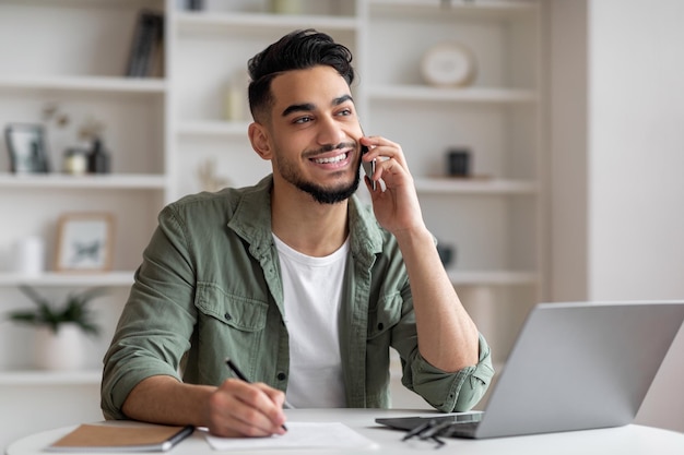 Smiling handsome millennial islamic guy manager with beard speaks by phone at workplace with laptop