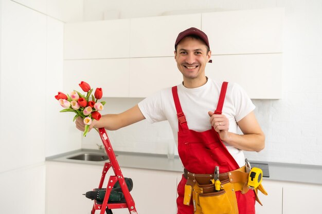 smiling handsome plumber holding bouquet flowers and looking at camera in kitchen.