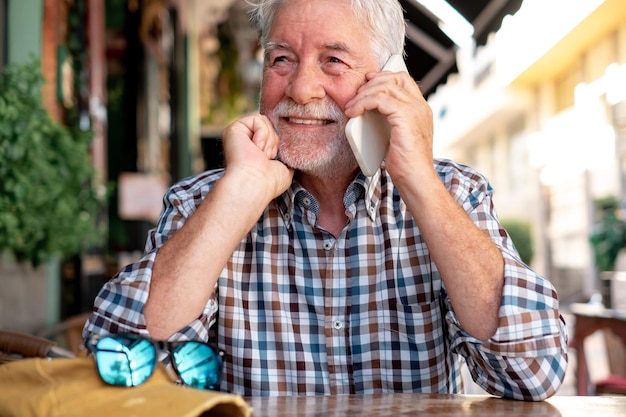 Smiling handsome senior man sitting outdoor at a cafe table\
holding mobile phone and talking caucasian elderly man relax in\
retirement or vacation