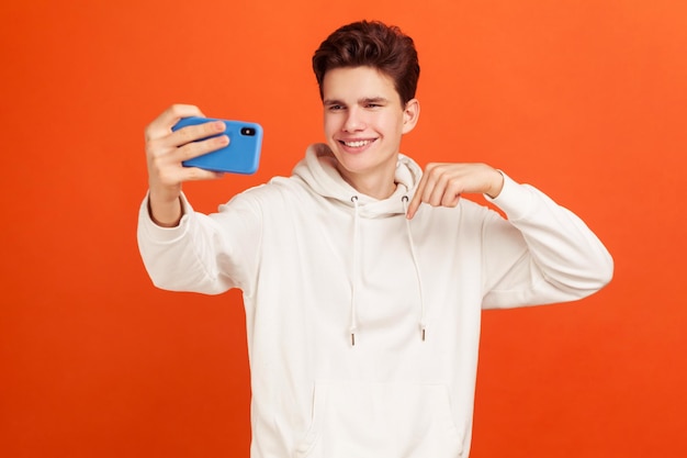 Smiling handsome teenager with stylish hairdo in casual hoodie posing in front of smartphone, pointing finger making selfie, blogging. Indoor studio shot isolated on orange background