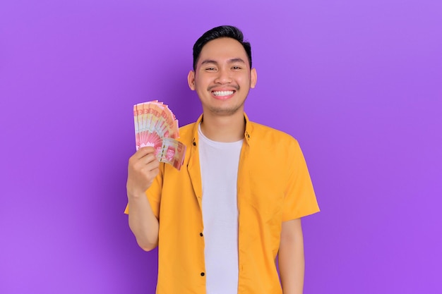 Smiling handsome young Asian man holding money banknotes isolated on purple background