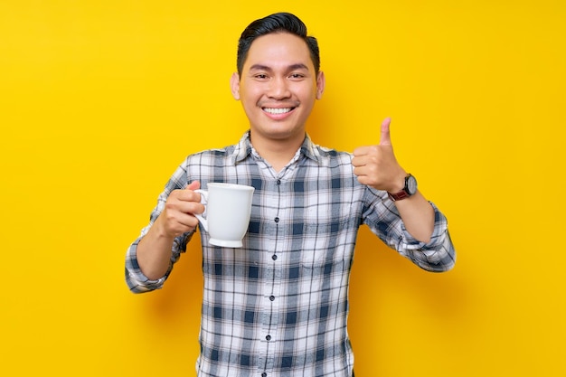 Smiling handsome young asian man wearing a white checkered shirt holding mug of coffee and showing thumbs up sign good taste isolated on yellow background people lifestyle concept