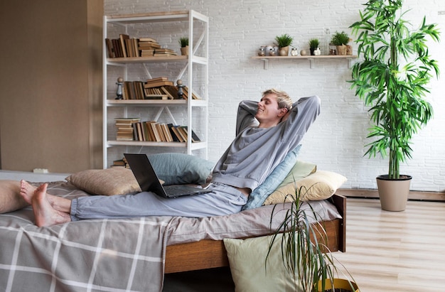 사진 웃고 있는 20세의 잘생긴 청년은 집에 있는 침실에서 시간을 보내며 침대에 누워 노트북 컴퓨터를 하고 있다