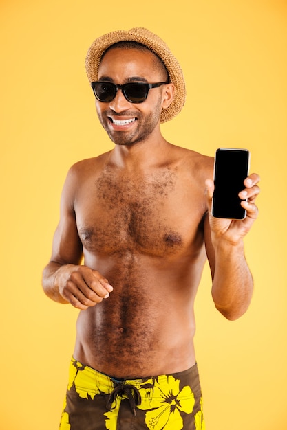 Улыбающийся красивый молодой человек в шляпе и солнцезащитных очках держит пустой экран смартфона над оранжевой стеной