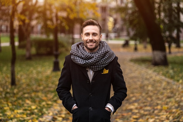 Smiling handsome young man looking at camera standing outdoors with hands in pockets of his dark blu...