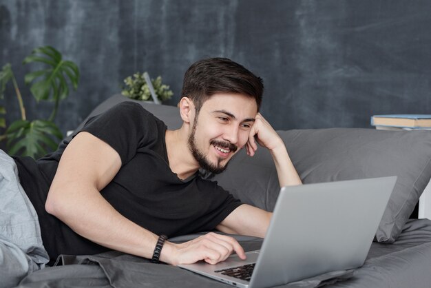 Smiling handsome young man lying in bed and using laptop while hatting with friends vie online app