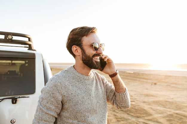 Smiling handsome young man using mobile phone while standing at the car at the sunny beach, calling