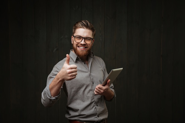 Smiling handsome young man using tablet and showing thumb up isolated on a black wooden surface