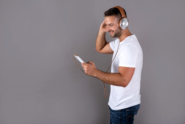 彼のヘッドフォンで音楽を聴き、灰色の上に立っている間彼のスマートフォンを保持している白いシャツを着た笑顔のハンサムな若い男